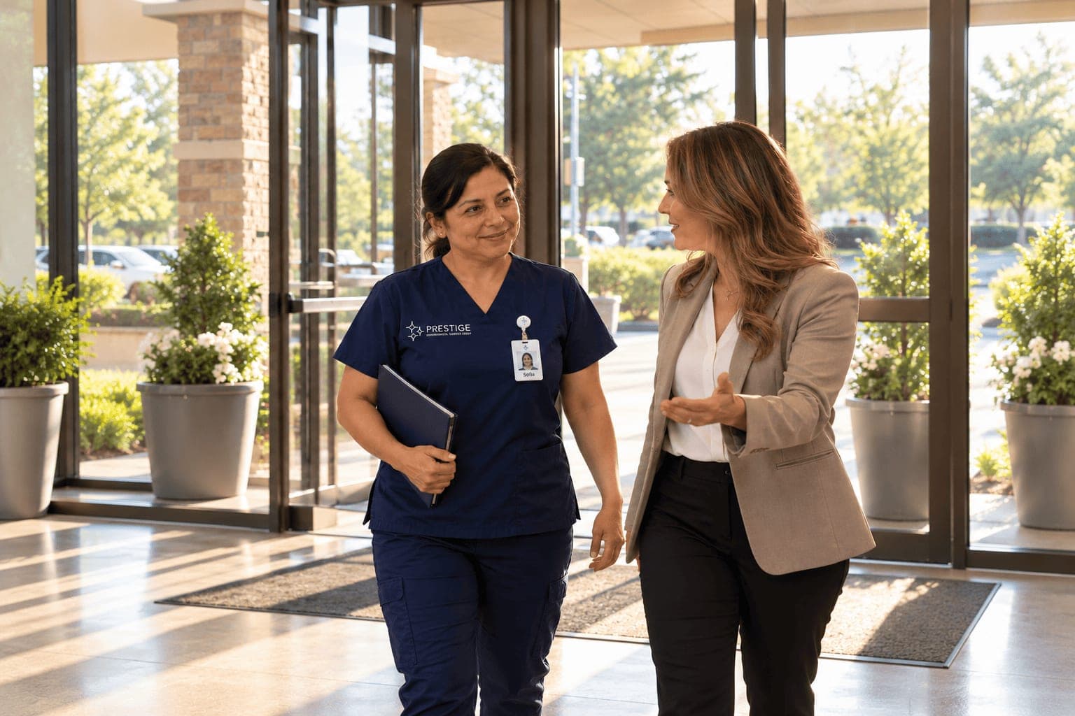 A Prestige housekeeping manager arriving at the main entrance of a facility with a folder under her arm, walking in beside the facility administrator.